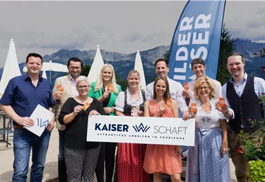 Eine Gruppe von Menschen steht zusammen und hält Getränke in den Händen. Im Hintergrund sind Berge und ein Werbebanner sichtbar.