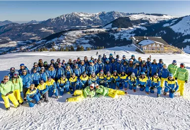 Eine Gruppe von Skifahrern steht auf einer schneebedeckten Piste. Im Hintergrund sind Berge und ein Aussichtspunkt zu sehen.