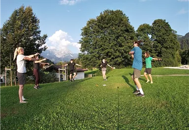 A group of six people is doing outdoor exercises on a green meadow. In the background, trees and mountains can be seen.