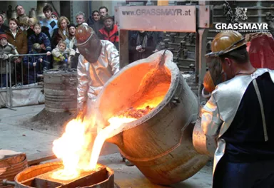 Two people in protective gear are pouring molten metal from a large cauldron. In the background, spectators are watching the spectacle.