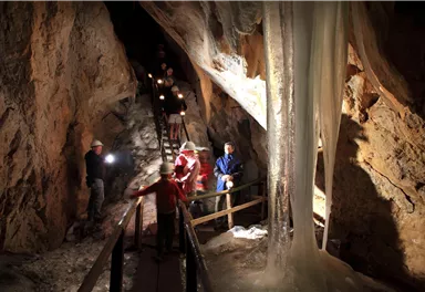 A group of people is exploring a cave with lighting. In the background, impressive rock formations and an ice waterfall can be seen.