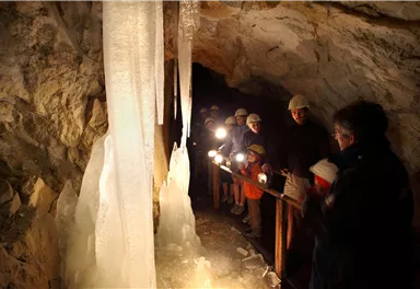 A group of people stands in a cave and observes ice formations. They are equipped with helmets and flashlights.