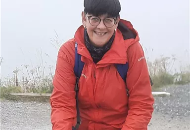 A person in a red jacket is sitting on a stone. Behind her is a misty background with plants.