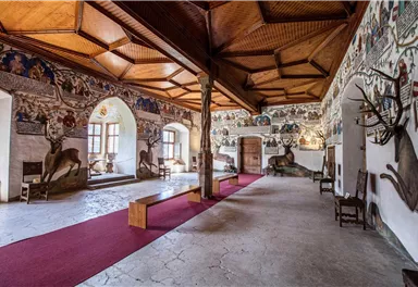 A historic room with an ornate wooden ceiling and walls adorned with animal motifs and paintings. The floor is covered with a red carpet and there are some wooden furniture pieces.