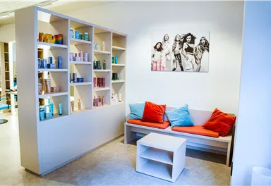 Ein moderner Empfangsbereich mit einem gemütlichen Sofa in orange und blau. Regale mit Pflegeprodukten sind an der Wand angebracht, und ein Bild von mehreren Frauen hängt im Hintergrund.