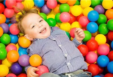 A smiling child is lying in a colorful ball pit. The colorful balls are in different colors such as yellow, blue, and red.