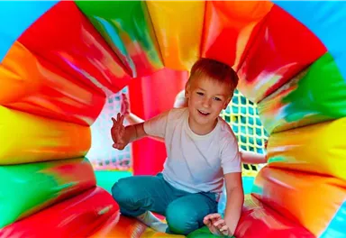 A cheerful boy is playing in a colorful jump tunnel. He is smiling and raising his hand in the air.
