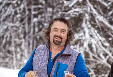 A smiling man in the snow is wearing a colorful jacket. The background is covered with snowy trees.