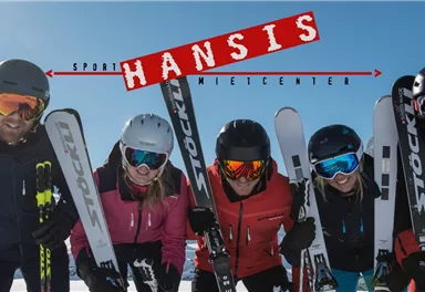 A group of five skiers smiles in the sun. In the background, a clear blue sky can be seen.