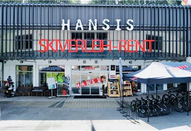 A shop with the sign "Hansis Skiverleih-Rent" and a large selection of sporting goods. Several bicycles for rent are standing in front of the shop.