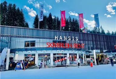 A modern ski shop with the sign "HANSIS SKIVERLEIH" in the midst of a snowy environment. In the background, trees and a blue sky can be seen.