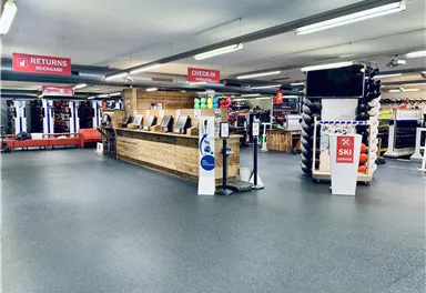 A modern sports store with a reception counter and various sports items. In the background, signs for returns and the checkout area are visible.
