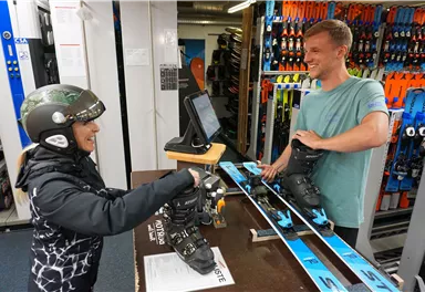 A female skier receives her equipment in a sports store. The staff member kindly helps her with putting on her ski boots.