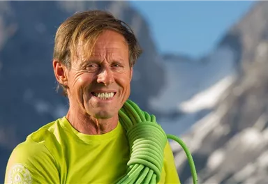 A smiling man is carrying a green climbing rope over his shoulder. Impressive mountains can be seen in the background.