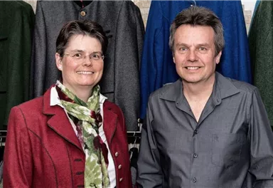 Ein Paar steht in einem Bekleidungsgeschäft vor einer Auswahl von Jacken. Die Person links trägt einen roten Blazer und einen Schal, während die Person rechts ein graues Hemd anhat.