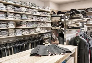 Ein gut sortierter Bekleidungsladen mit vielen übereinandergestapelten Hemden und Kleidungsstücken. Auf einem Tisch liegt ein hochwertiger Anzug zur Ansicht.