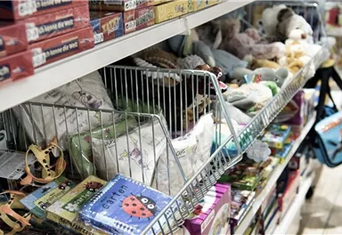 Ein Regal mit Spielzeugen und Büchern, das viele verschiedene Artikel zeigt. Auf der linken Seite sind Puzzle und Stofftiere, während rechts ein buntes Torba zu sehen ist.