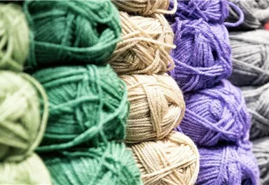 Eine farbenfrohe Sammlung von Wollknäueln in verschiedenen Farben. Die Wolle ist ordentlich gestapelt und zeigt grüne, beige, lila, graue und rote Töne.