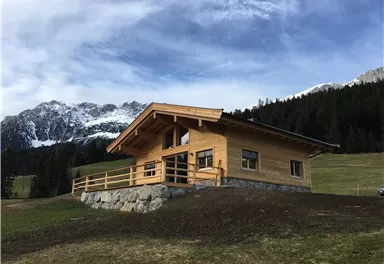 Ein modernes Holzhaus in einer malerischen Umgebung. Im Hintergrund sind die Berge und ein klarer Himmel zu sehen.