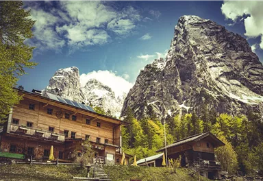 Eine malerische Berglandschaft mit hohen Gipfeln und einer charmanten Holzunterkunft im Vordergrund. Der Himmel ist teilweise bewölkt und die Natur ist üppig grün.