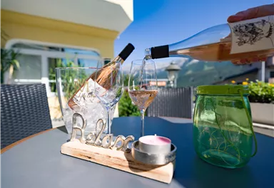 Ein Tisch auf einer Terrasse mit einer Flasche Roséwein, die in ein Glas eingeschenkt wird. Im Hintergrund sind Pflanzen und ein klarer Himmel sichtbar.