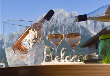 Ein elegantes Getränkeset mit einer Flasche Roséwein und zwei Gläsern steht auf einem Holztisch. Im Hintergrund sind Berge und ein klarer Himmel zu sehen.