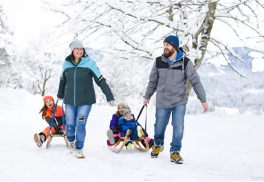 A family is taking a walk in the snow. The children are sitting on sleds while the parents are pulling them.