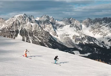 Two skiers are descending a snowy slope, surrounded by majestic mountains. The sky is partly cloudy and the landscape is wintery beautiful.
