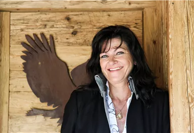 A woman smiles in front of a wooden wall with an eagle emblem. She is wearing an elegant jacket and appears friendly and inviting.
