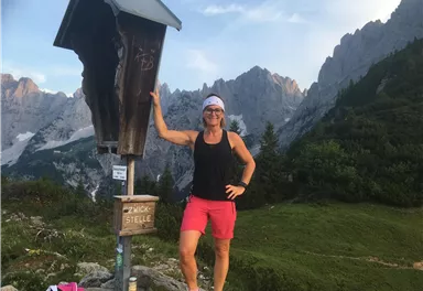 A woman is standing in front of a hiking signpost in the mountains. Majestic mountain peaks and a green landscape can be seen in the background.