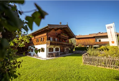 A traditional wooden house with a well-maintained garden and blue sky. In the background, some modern buildings can be seen.