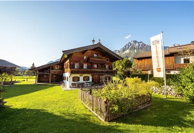 A traditional alpine wooden house with a lovingly designed garden. In the background, majestic mountains and a clear blue sky can be seen.