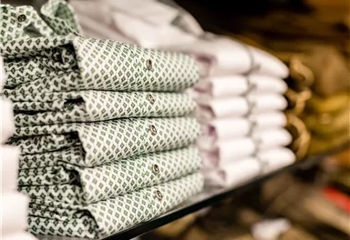 Gestapelte Hemden, einige mit grünem Muster, lagern auf einem Regal. Im Hintergrund sind weitere faltbare Hemden sichtbar.