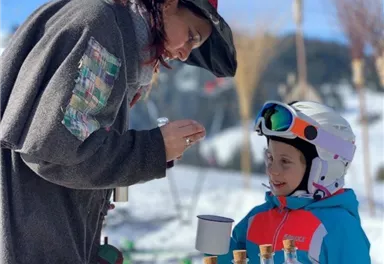A witch is showing a child various colorful liquids from jars on a table in the snow. In the background, there is a snow-covered landscape and a blue sky.