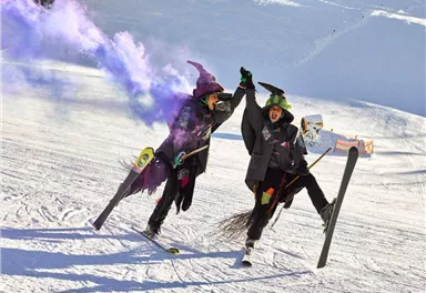 Two people on skis are wearing witch costumes and are celebrating joyfully. They are raising their hands high and letting purple smoke rise.