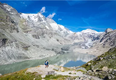 Eine atemberaubende Berglandschaft mit schneebedeckten Gipfeln und einem klaren See. Zwei Wanderer genießen die Aussicht in der Natur.