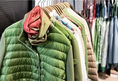 Eine Auswahl an modischer Kleidung, darunter eine grüne Jacke und verschiedene Oberteile. Die Kleidungsstücke hängen ordentlich auf einer Stange in einem Geschäft.