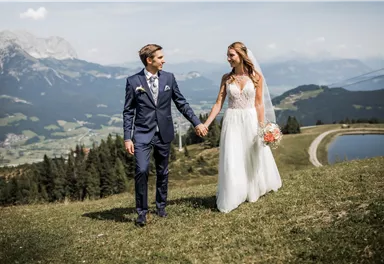 A bride and groom stand on a meadow in the mountains. In the background, green hills and a blue sky can be seen.
