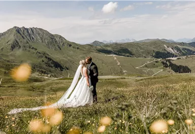A bride and groom kiss in a picturesque mountain landscape. The meadow is adorned with colorful flowers, and the mountains provide a stunning backdrop.