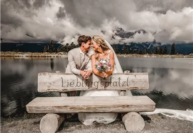 A loving couple is sitting on a bench by the water. In the background, dark clouds and mountains can be seen.