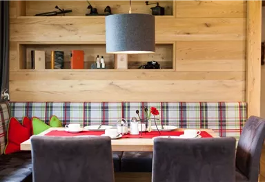 Ein einladender Essbereich mit einem holzverkleideten Hintergrund. Der Tisch ist liebevoll gedeckt und mit farbigen Kissen sowie einer Blumendekoration ausgestattet.