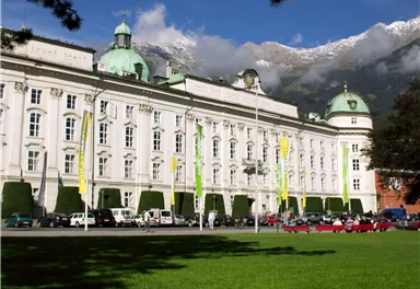 An impressive building with green domes and a beautiful facade. In the foreground, there is a green meadow, and the surrounding mountains are partially covered with snow.