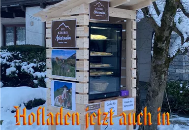 Ein kleiner Verkaufsstand im Freien für lokale Produkte steht in einer verschneiten Umgebung. Auf dem Stand sind Schilder mit dem Namen "Käserei Ackerhube" und Informationen über das Angebot zu sehen.