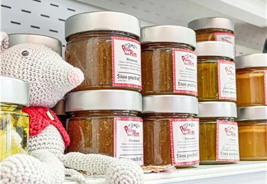 A shelf with various jars containing different jams or spreads. Next to the jars sits a cozy-looking, crocheted soft toy.