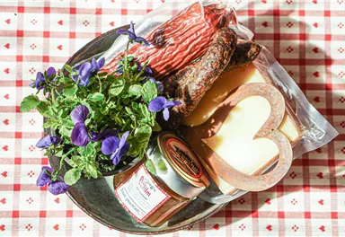 A rustic plate with sausage, cheese, and jam. In addition, a small pot with purple flowers.