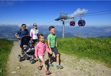 Eine Familie genießt einen sonnigen Tag in den Bergen. Im Hintergrund sieht man eine Seilbahn und eine blühende Landschaft.