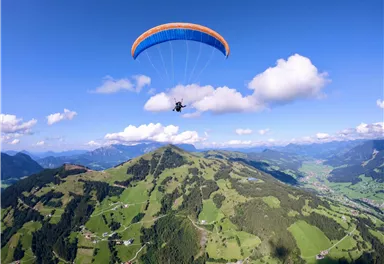 A paraglider floats above a green mountain landscape with blue sky and white clouds. Below, picturesque villages and meadows are visible.