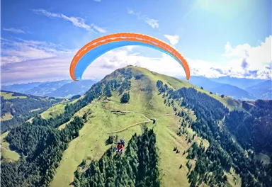 A mountainous region with green meadows and dense forests. A paraglider flies over the landscape in the bright blue sky.