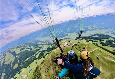 A couple paragliding over green hills and beautiful scenery. The sky is blue with some clouds.