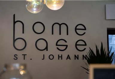 A modern sign with the inscription "home base St. Johann" on a wall. In the background, a plant and a warm light source can be seen.